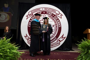 This image depicts a man presenting an award to a woman at a graduation ceremony. Both people are in graduation regalia. The stage behind them has the College of the Ozarks logo. There are two plants on either side of them.  